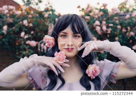 Fairy tale woman portrait. Young beautiful woman Fairy holds pink flower rose on blooming bush background. Hairstyle loose green hair, romantic purple medieval vintage dress. Summer fantasy garden. 115272344