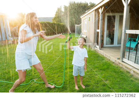Joyful kid with mother playing with water sprinkler hose, splashing, laughing, having fun together in garden on sunset. Summer outdoor activity. Happy active childhood, family time. Selective focus. 115272362