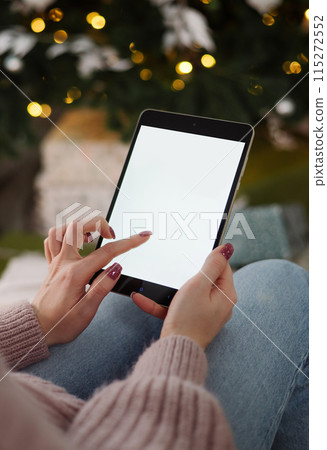 Mock-up, tablet in the hands of a woman. Christmas tree background. Concept of online shopping, discounts Mock-up, tablet in the hands of a woman. Christmas tree background. Concept of online shopping, discounts 115272552