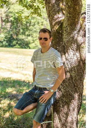 Portret of a 40 yo man, standing relaxed against a tree in summer, Kordel,Germany 115273188