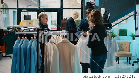 Fashion stylist suggesting clothing items to regular customer, team of store workers help elderly woman to buy trendy clothes on sale. Retail clerks assisting client during shopping spree. Camera B. 115275215