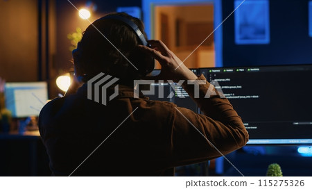 Engineer wearing headphones while using coding to fortify security, remotely working for IT company. Software expert listening music and upgrading binary code scripts on computer, camera A 115275326