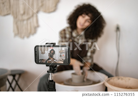 Young woman potter sculpting jug on potter's wheel while recording vlog Young woman potter sculpting jug on potter's wheel while recording vlog 115276355