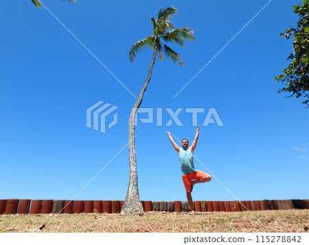Man Does Tree Pose with Arms raised into the air at beach park 115278842