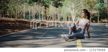 Young woman in athletic wear sitting on a forest road, resting after a workout, with trees and sunlight in the background. Young woman in athletic wear sitting on a forest road, resting after a workout, with trees and sunlight in the background. 115279489