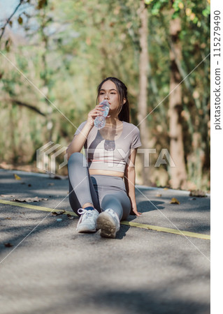 A young woman in sportswear sits on a pathway in a forest, drinking water and taking a break from her workout on a sunny day. A young woman in sportswear sits on a pathway in a forest, drinking water and taking a break from her workout on a sunny day. 115279490