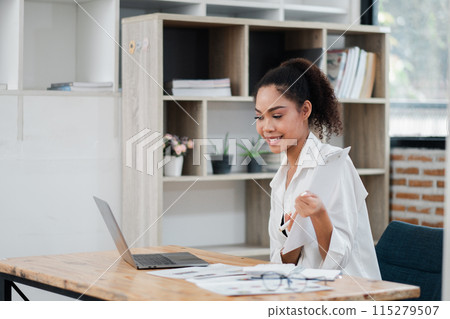 A woman is sitting at a desk with a laptop and a stack of papers. She is smiling and holding a piece of paper. The scene suggests a positive and productive work environment 115279507