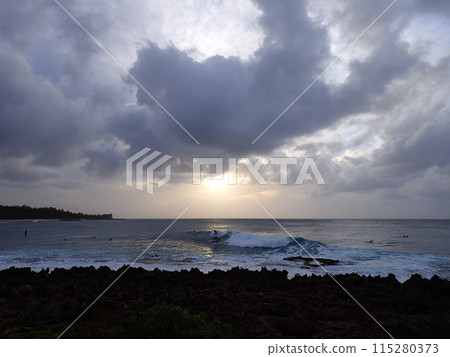 Surfers ride waves as sunsets though the clouds off the coast of Turtle Bay Surfers ride waves as sunsets though the clouds off the coast of Turtle Bay 115280373