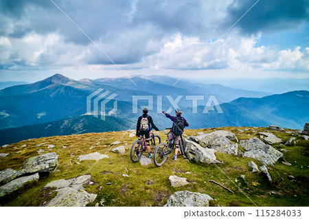 Two cyclists men riding electric bikes outdoors. Back view of male tourists resting on the top of hill, enjoying beautiful mountain landscape, wearing helmet and backpack. Concept of active leisure. 115284033