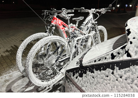 Electric vehicle and bicycles at self-service car wash during nighttime, covered in foam, indicating that it is in the middle of a wash cycle. Bicycles mounted on a car rack at the back. Electric vehicle and bicycles at self-service car wash during nighttime, covered in foam, indicating that it is in the middle of a wash cycle. Bicycles mounted on a car rack at the back. 115284038