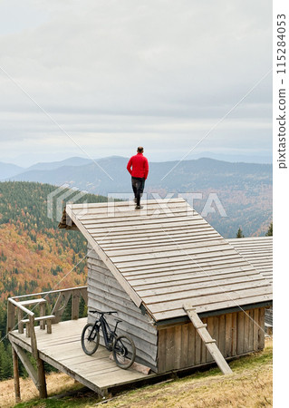 Person resting on edge of steeply sloped wooden rooftop in mountainous area after riding on electric mountain bike on cloudy day. Below the rooftop, there is wooden deck with mountain bicycle. Person resting on edge of steeply sloped wooden rooftop in mountainous area after riding on electric mountain bike on cloudy day. Below the rooftop, there is wooden deck with mountain bicycle. 115284053