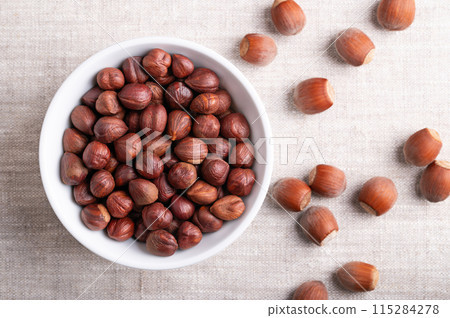 Hazelnuts in a white bowl on linen fabric. Whole, dried and shelled nuts of Corylus avellana, on the right side in the shells. Ready to eat as snack, and also used for baking. Close-up, from above. 115284278