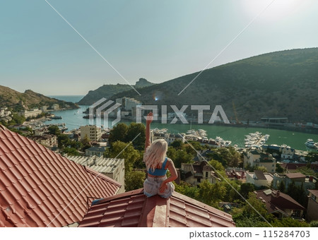 Woman sits on rooftop, enjoys town view and sea mountains. Peaceful rooftop relaxation. Below her, there is a town with several boats visible in the water. Rooftop vantage point. Woman sits on rooftop, enjoys town view and sea mountains. Peaceful rooftop relaxation. Below her, there is a town with several boats visible in the water. Rooftop vantage point. 115284703