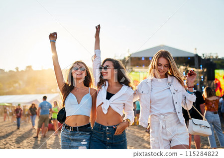 Young women dance at sunset on sandy shore, enjoying music festival. Friends celebrate, summer beach party vibe, arms uplifted in joy. Casual fashion, sea backdrop, golden hour light at music event. Young women dance at sunset on sandy shore, enjoying music festival. Friends celebrate, summer beach party vibe, arms uplifted in joy. Casual fashion, sea backdrop, golden hour light at music event. 115284822
