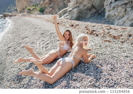 Two women are laying on the beach, one of them is wearing a bikini Two women are laying on the beach, one of them is wearing a bikini 115284856