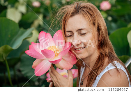 A woman is holding a pink lotus flower and smiling. The flower is surrounded by green leaves, and the woman is standing in a field of flowers. 115284937
