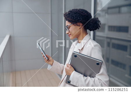 Busy female entrepreneur with laptop is using phone while standing on office terrace  115286251