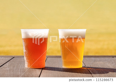 桌子上有兩種啤酒。使用塑膠杯。麥田背景 115286673