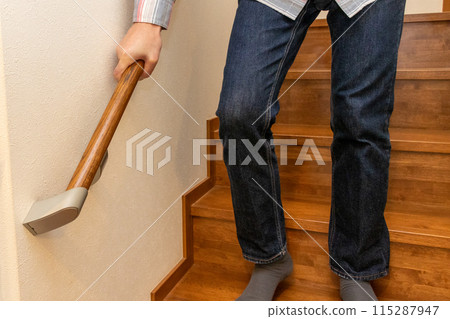 A person carefully descends the stairs, holding on to the banister A person carefully descends the stairs, holding on to the banister 115287947