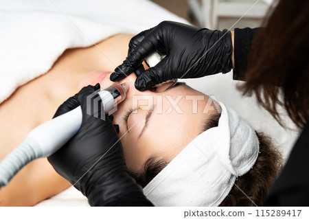 A close-up cosmetologist manipulates a hydropeeling machine to clean and rejuvenate a patient's skin in a modern clinic. 115289417