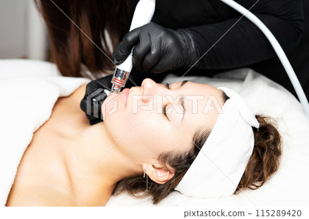 A close-up cosmetologist manipulates a hydropeeling machine to clean and rejuvenate a patient's skin in a modern clinic. 115289420