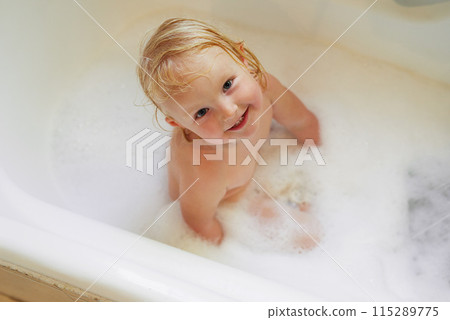 Portrait, bathtub and top view of happy baby girl in water for hygiene, health or wellness in bathroom. Foam, soap and face of kid bathing, clean skin and cute infant child with bubbles in home above 115289775