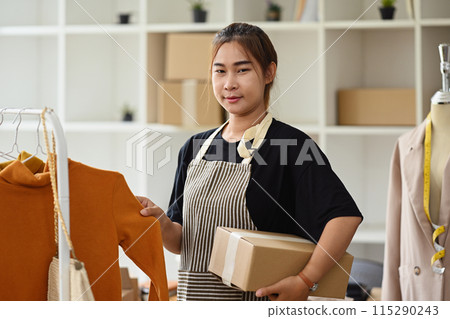 Portrait of young female small business owner holding cardboard box standing at her clothing shop 115290243
