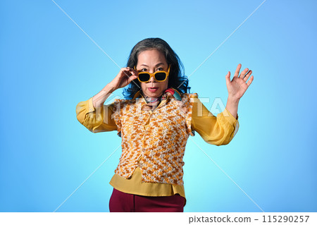 Attractive mature woman with fashionable clothes and sunglasses posing over blue background 115290257