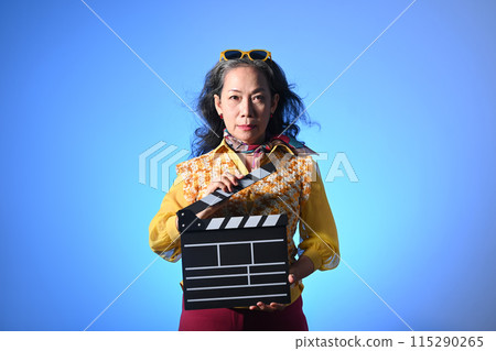 Fashionable middle age woman holding clapperboard isolated on blue background 115290265