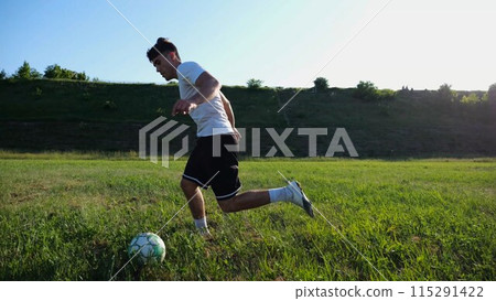 Professional footballer in uniform shows tricks with ball while running at green field. Sportsman kicks ball during jogging at meadow on sunny day. Young man training at stadium. Freestyle football 115291422