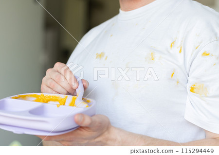 Father ended to feed his child. Father in a white tshirt messy all over after feeding his baby holding a plate with baby puree leftovers. 115294449