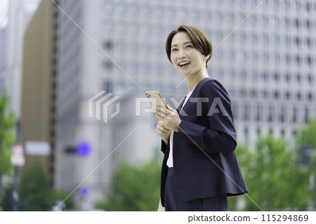 A businesswoman standing in an office district holding a smartphone 115294869