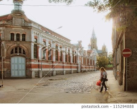 A woman with a red bag walks on a quiet, cobblestone street lined with historic brick buildings and a distant church tower at sunset. 115294888