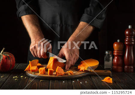 A man cuts a pumpkin into cubes with a kitchen knife on a wooden board. Fresh raw chopped pumpkin pieces in the kitchen. Preparing ingredients for a seasonal fall dish, soup or pie A man cuts a pumpkin into cubes with a kitchen knife on a wooden board. Fresh raw chopped pumpkin pieces in the kitchen. Preparing ingredients for a seasonal fall dish, soup or pie 115295724