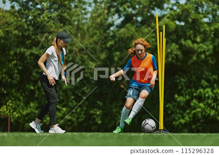 Dynamic Sports Coaching. Girl in colorful soccer uniform demonstrating her dribbling skills, playing football with guidance of female coach. Dynamic Sports Coaching. Girl in colorful soccer uniform demonstrating her dribbling skills, playing football with guidance of female coach. 115296328