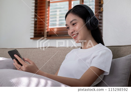 Woman wearing headphones relaxing on sofa using smartphone. Concept of digital leisure and entertainment Woman wearing headphones relaxing on sofa using smartphone. Concept of digital leisure and entertainment 115298592