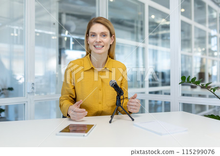 Woman in a yellow shirt leading a virtual meeting with a microphone and a tablet in a bright, modern office 115299076