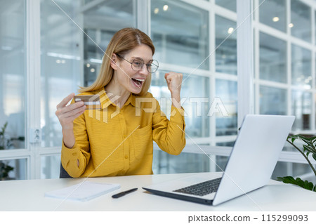 Excited woman in an office holding a credit card while using a laptop for online shopping. Celebrating successful purchase. 115299093