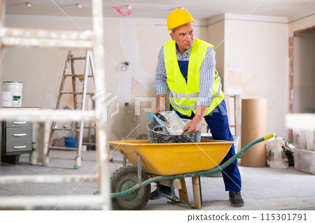 Worker in yellow vest and a protective helmet transports painting tools in wheelbarrow Worker in yellow vest and a protective helmet transports painting tools in wheelbarrow 115301791
