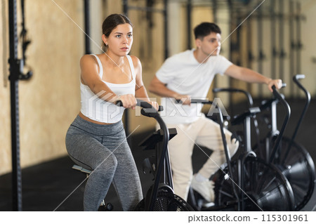 Positive girl riding exercise airbike for cardio workout during cycling class at modern crossfit gym Positive girl riding exercise airbike for cardio workout during cycling class at modern crossfit gym 115301961