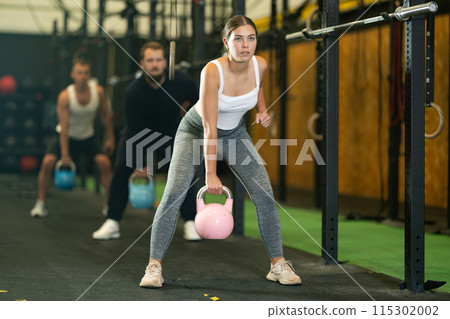 Sportive young woman lifting heavy kettlebell in sports hall 115302002