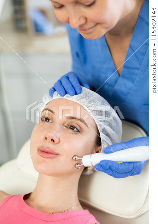 Adult woman cosmetologist doing microcurrent procedure to young woman 115302143