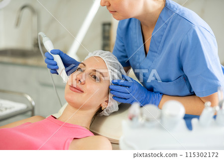 Caucasian brunette young girl lying in spa while having facial treatment procedures with modern technologies vibrating shovel leather knife. Soften skin keratin, deep clear skin dirt. Hardware Caucasian brunette young girl lying in spa while having facial treatment procedures with modern technologies vibrating shovel leather knife. Soften skin keratin, deep clear skin dirt. Hardware 115302172