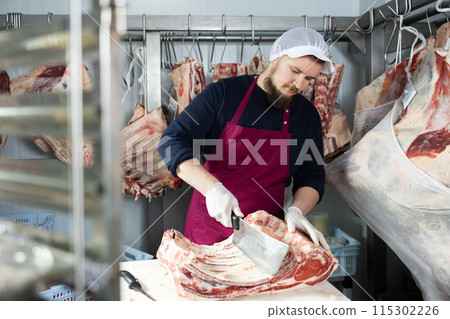 Bearded butcher cutting beef ribs in cold storage room of store 115302226