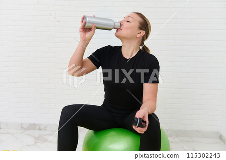 Young sporty female athlete hydrating while exercising with gym equipment at home Young sporty female athlete hydrating while exercising with gym equipment at home 115302243