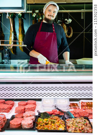 Smiling butcher behind counter with meat products in refrigerated showcase Smiling butcher behind counter with meat products in refrigerated showcase 115302358