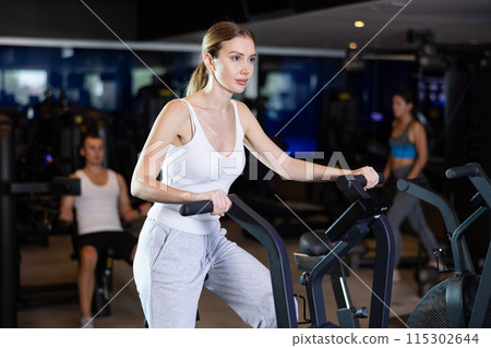 Girl performs exercises with stationary air bicycle in gym Girl performs exercises with stationary air bicycle in gym 115302644