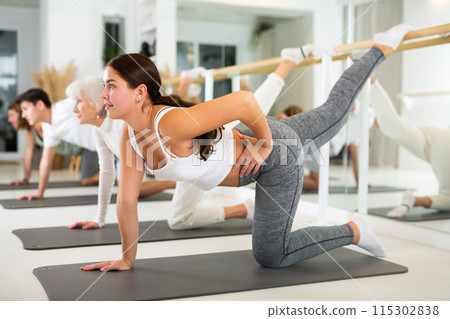 Positive young female performing set of exercises on a pilates mat during group class in fitness studio. 115302838