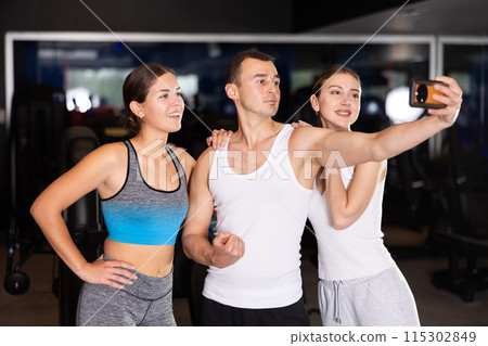 Two cheerful girls and young man taking selfie in fitness hall Two cheerful girls and young man taking selfie in fitness hall 115302849