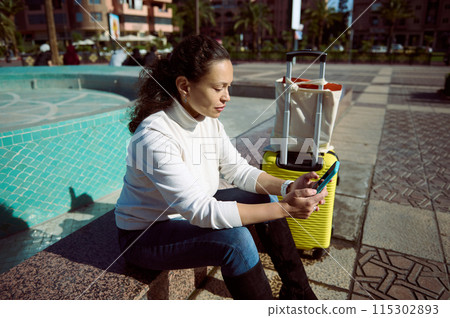 Woman sitting by a fountain with luggage, using smartphone outdoors Woman sitting by a fountain with luggage, using smartphone outdoors 115302893